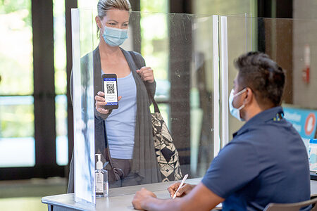 A middle aged woman enters a community centre and shows the attendant her vaccine passport on her cell phone. She is dressed casually with her purse over her shoulder. Both the woman and the attendant are wearing medical masks to protect them from COVID.
