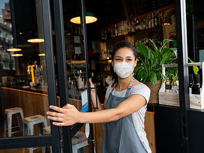 Happy business owner opening the door at a cafe wearing a facemask to avoid the spread of coronavirus â€“ reopening after COVID-19 concepts