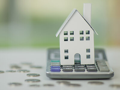 A white metal toy house sits on a calculator and is surrounded by coins. Illustrating the concepts of cost and expenses of real estate and buying a new home.