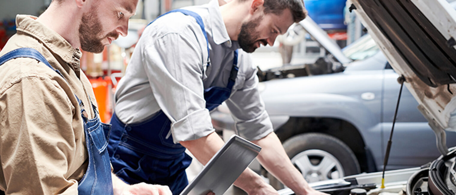Zwei Kfz-Mechatroniker reparieren den Motor eines Pkws bei aufgeklappter Motorhaube. Check mit Laptop,