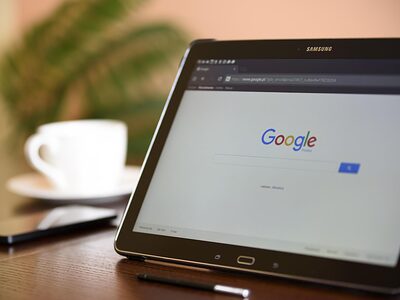 Google-Startseite auf einem Tablet. Im Hintergrund ein Smartphone und eine Kaffeetasse.