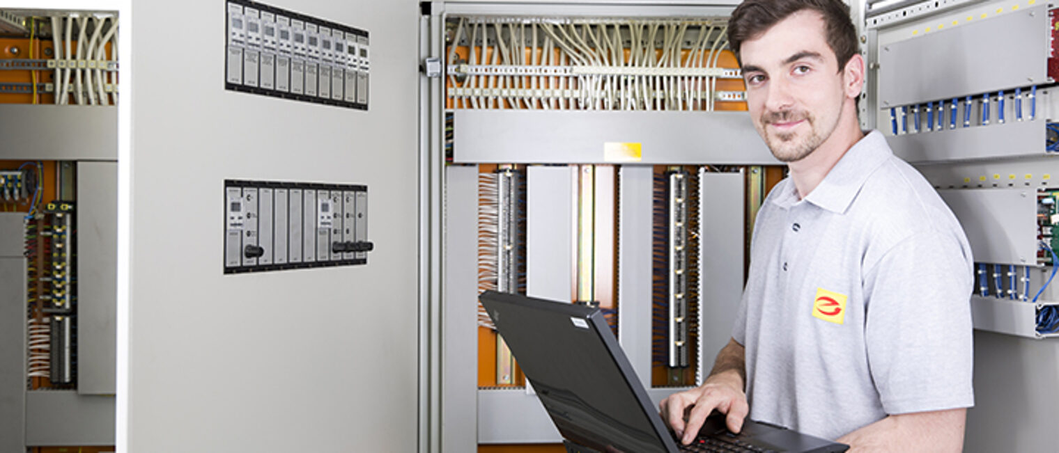 Ein Elektroniker für Automatisierungstechnik steht mit einem Laptop vor einem Schaltschrank, mit Blick in die Kamera Ein Elektroniker für Automatisierungstechnik steht mit einem Laptop vor einem Schaltschrank, mit Blick in die Kamera