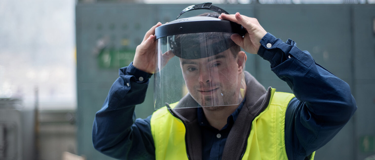 Mann mit Down-Syndrom trägt in der Werkstatt Schutzhelm und Warnweste. Mann mit Down-Syndrom trägt in der Werkstatt Schutzhelm und Warnweste.