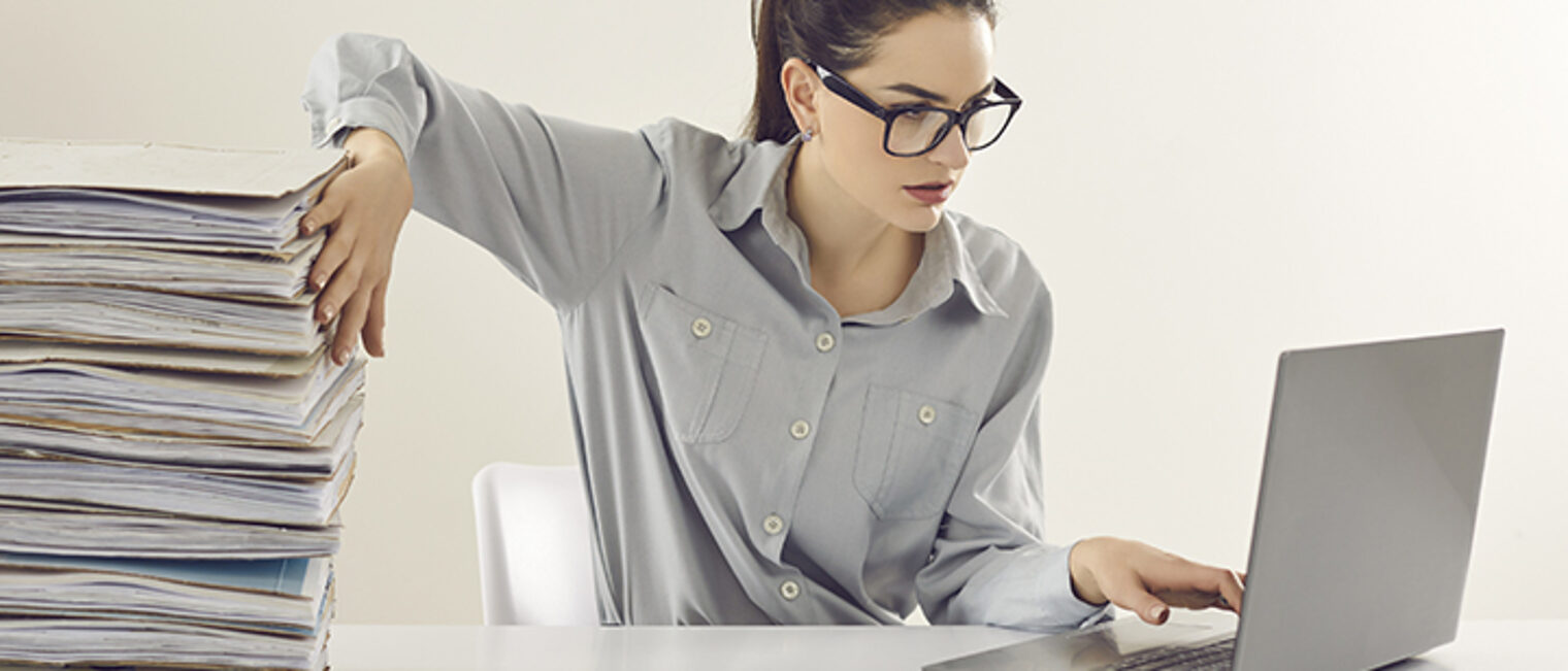 Eine Frau sitzt am Laptop und schiebt einen Stapel Papier beiseite. Eine Frau sitzt am Laptop und schiebt einen Stapel Papier beiseite.