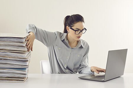 Eine Frau sitzt am Laptop und schiebt einen Stapel Papier beiseite.