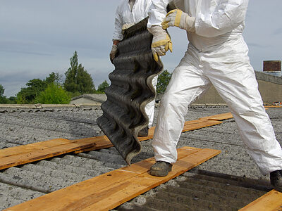 Zwei Männer in Schutzanzügen demontieren asbesthaltige Dachplatten