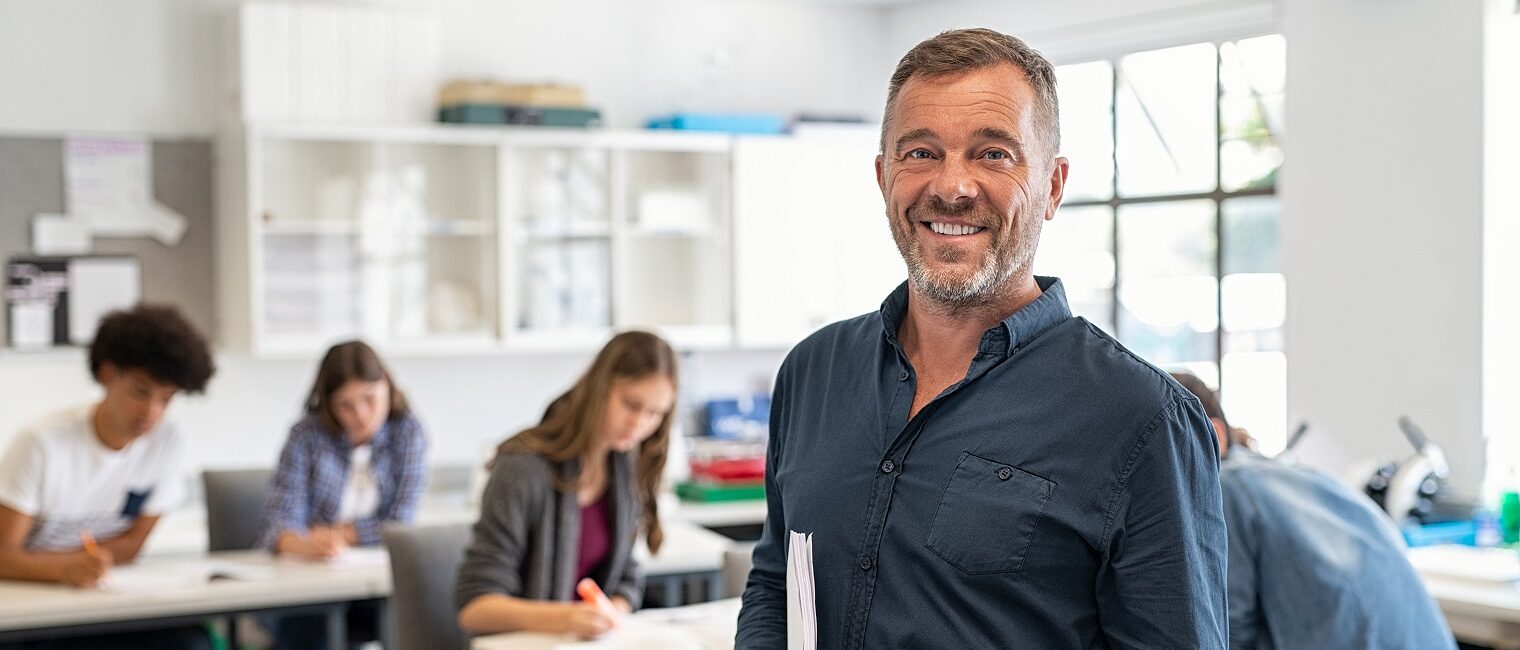 Portrait of mature teacher standing in university library and looking at camera with copy space. Happy mid adult lecturer at classroom standing after giving lecture. Satisfied high school teacher smiling and looking at camera while his studets studying in background.