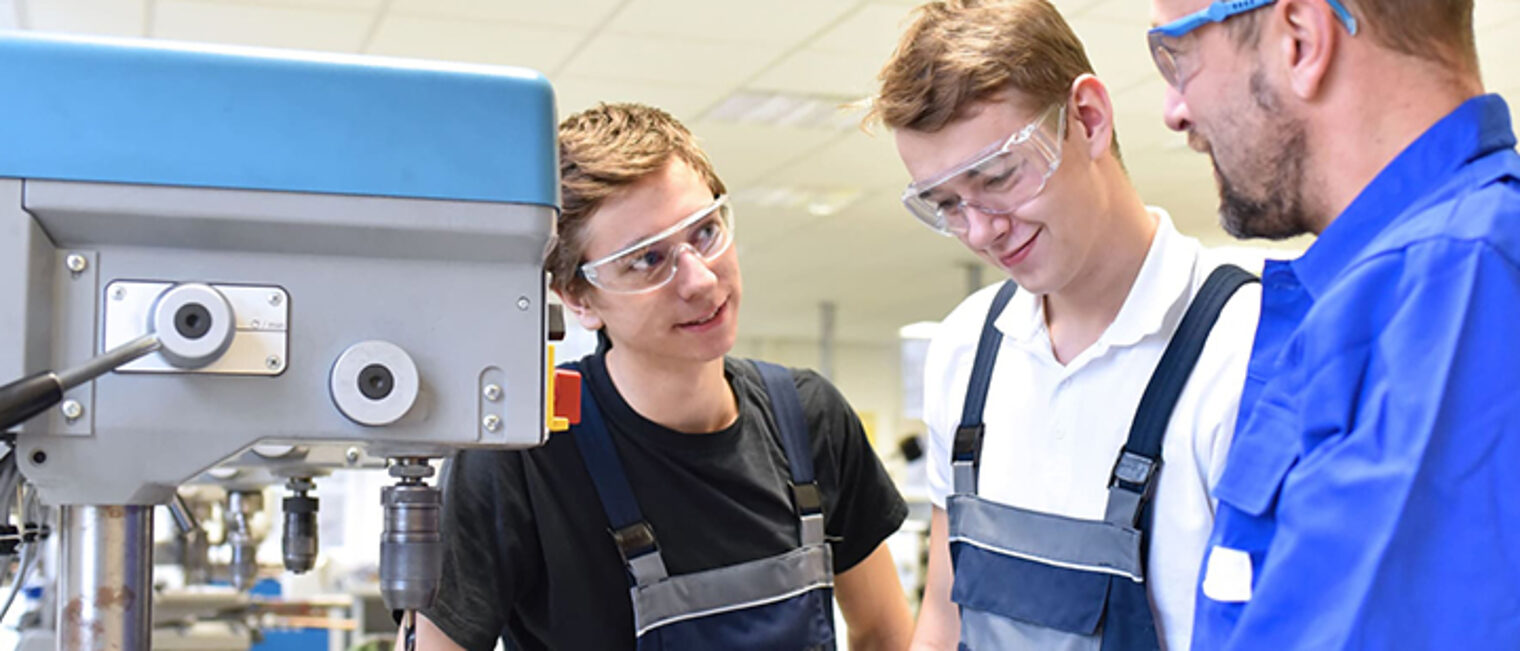 Zwei junge Männer mit Schutzbrille und einem Praktikumsanleiter stehen an einer Bohrmaschine. Zwei junge Männer mit Schutzbrille und einem Praktikumsanleiter stehen an einer Bohrmaschine.