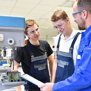 Zwei junge Männer mit Schutzbrille und einem Praktikumsanleiter stehen an einer Bohrmaschine.