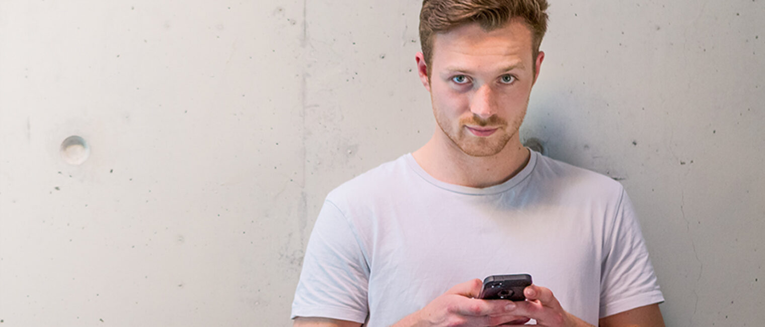 Junger Mann im T-Shirt mit Smartphone vor weißer Wand