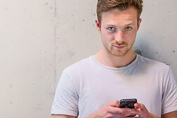 Junger Mann im T-Shirt mit Smartphone vor weißer Wand