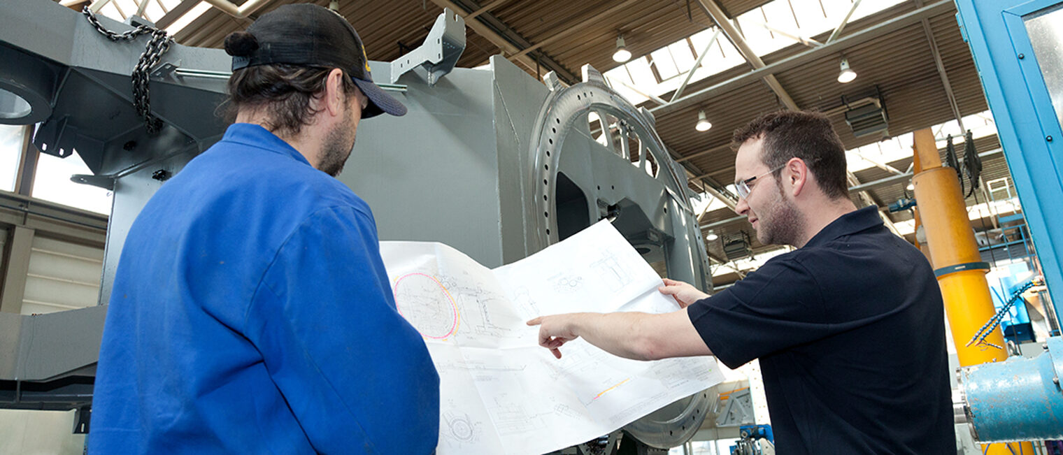 Zwei Männer blicken in einer Werkhalle auf einen Plan.