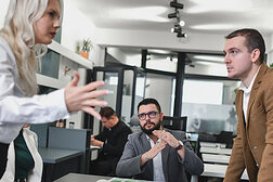 Zwei streitende Personen in einem Meeting, eine dritte beschwichtigt mit einem Handzeichen.