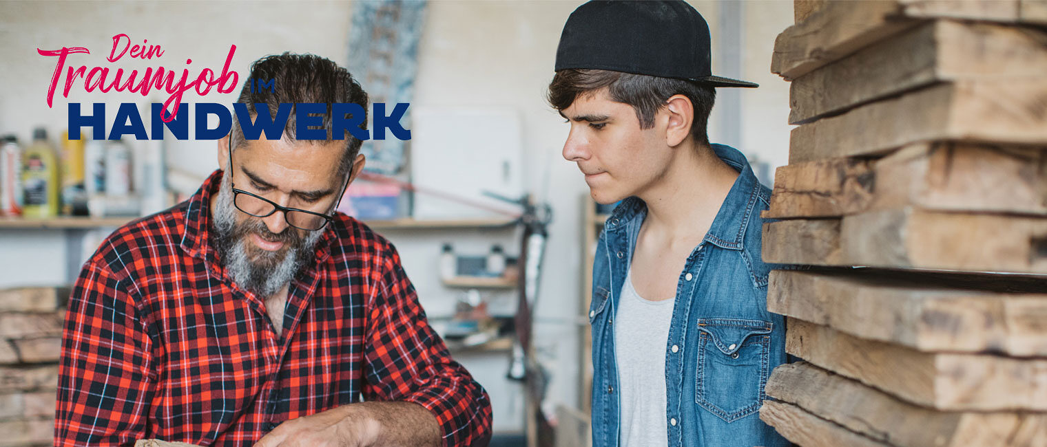 Handwerker in Tischlerei erklärt jungem Auszubildendem verschiedene Holzarten