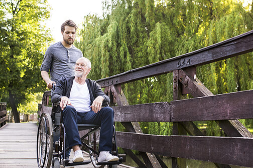 Junger Mann schiebt älteren Mann im Rollstuhl über eine Brücke.
